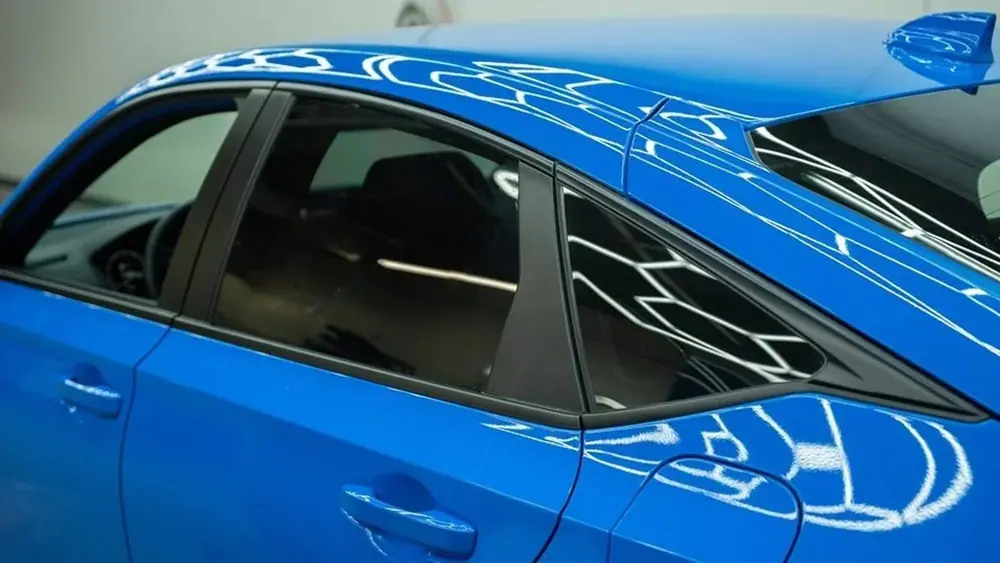 Close-up of freshly tinted rear windows on a bright blue sedan, hexagon shop lights reflecting in the paint
