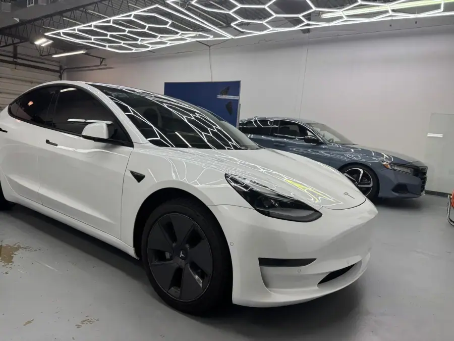 White Tesla Model 3 with dark tinted glass beside a blue sedan in a shop