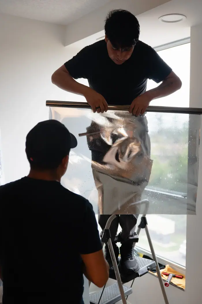 Two installers laying a sheet of window film on a tall pane, one on a step ladder